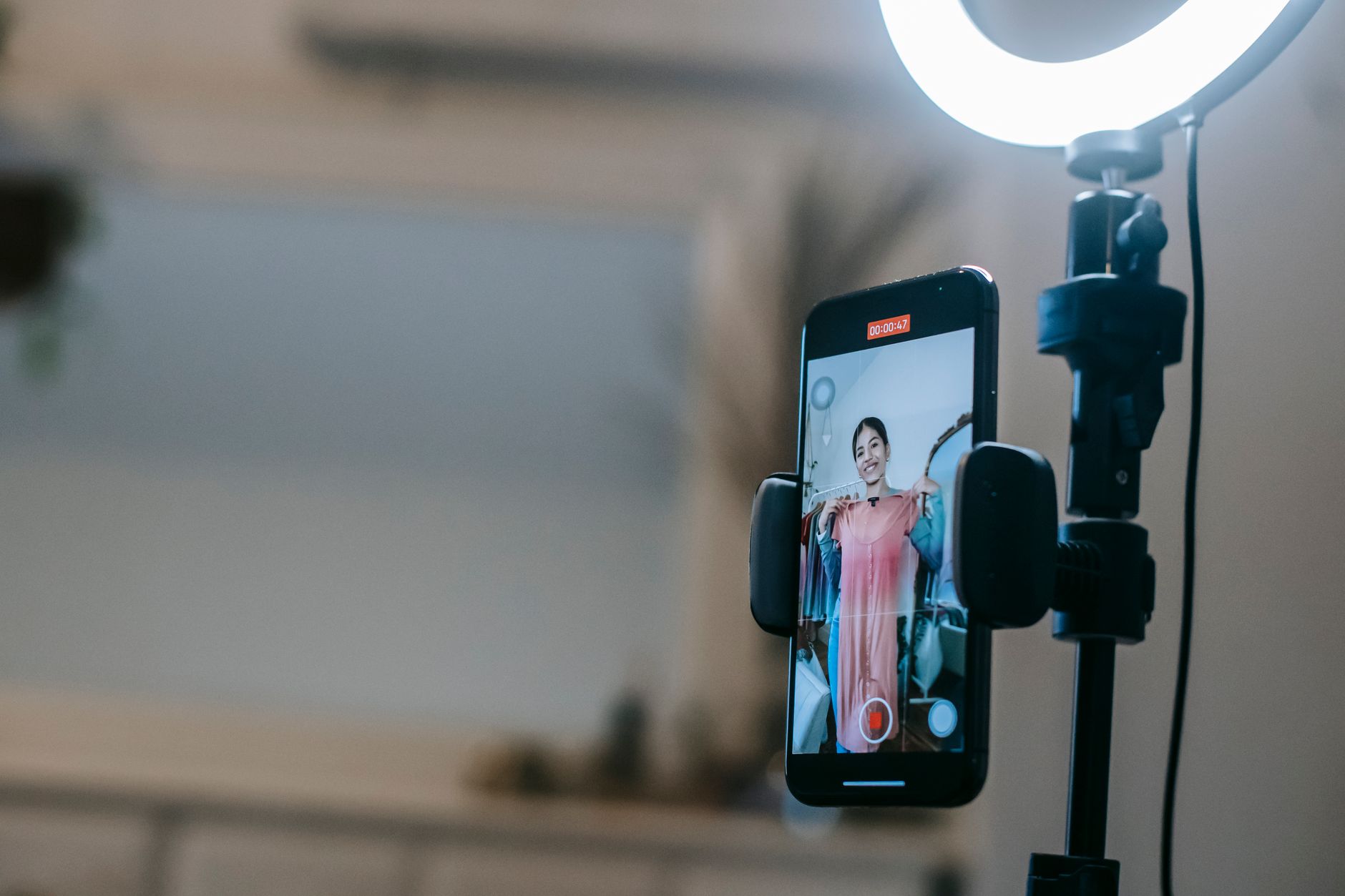 A smartphone clamped to a ring-light stand, recording a woman who holds a pink dress up to the camera