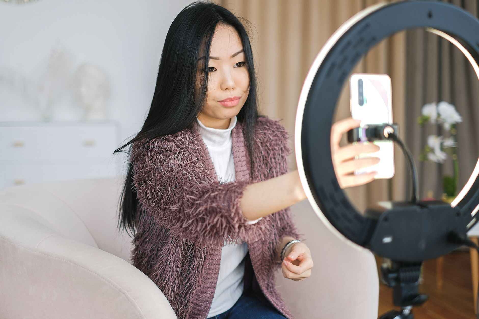 A woman seated on a sofa reaching out to adjust a smartphone held in a ring light