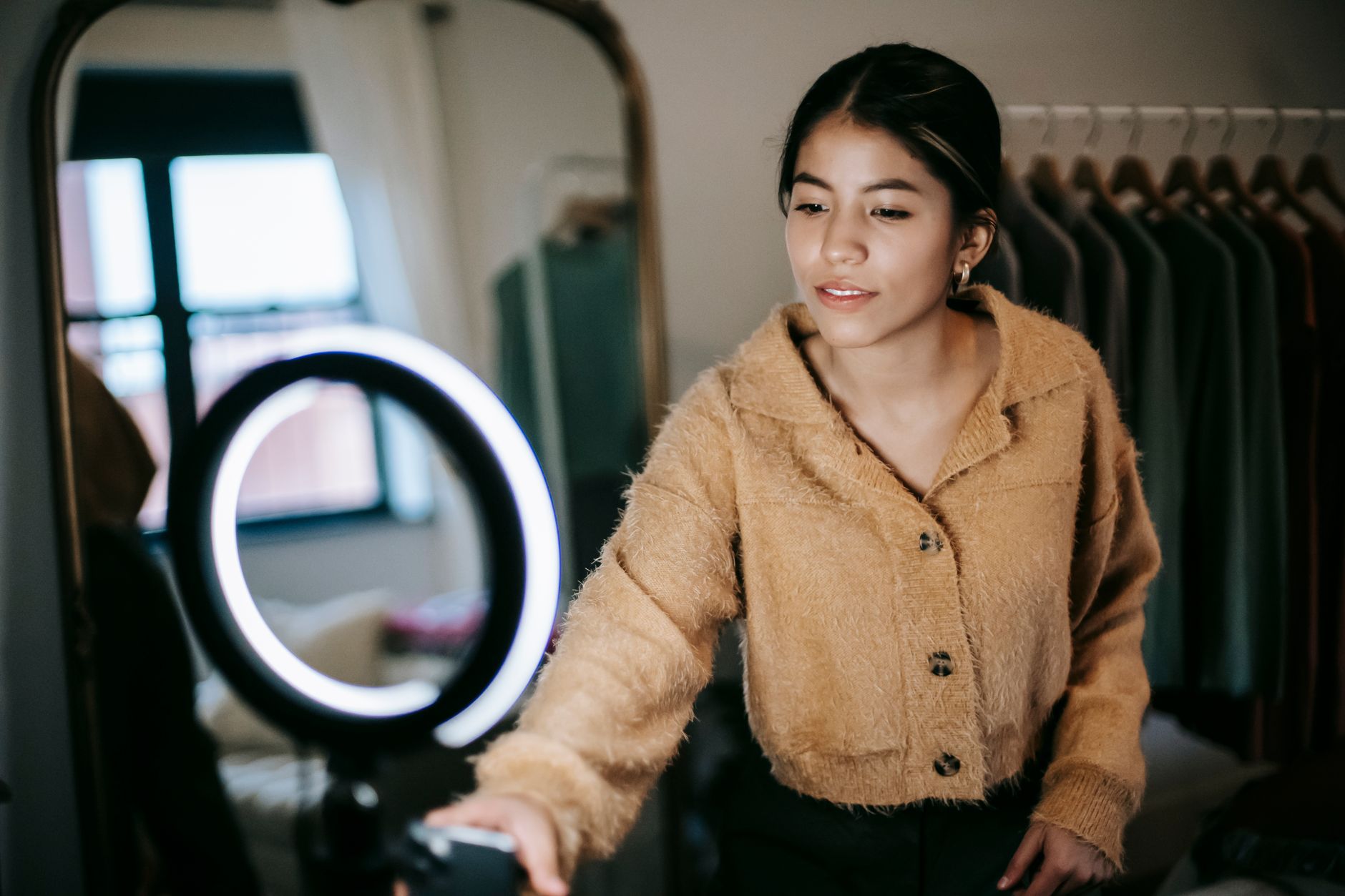 A woman in a knitted cardigan adjusting a ring light in a bedroom, with a mirror and a clothes rail behind her