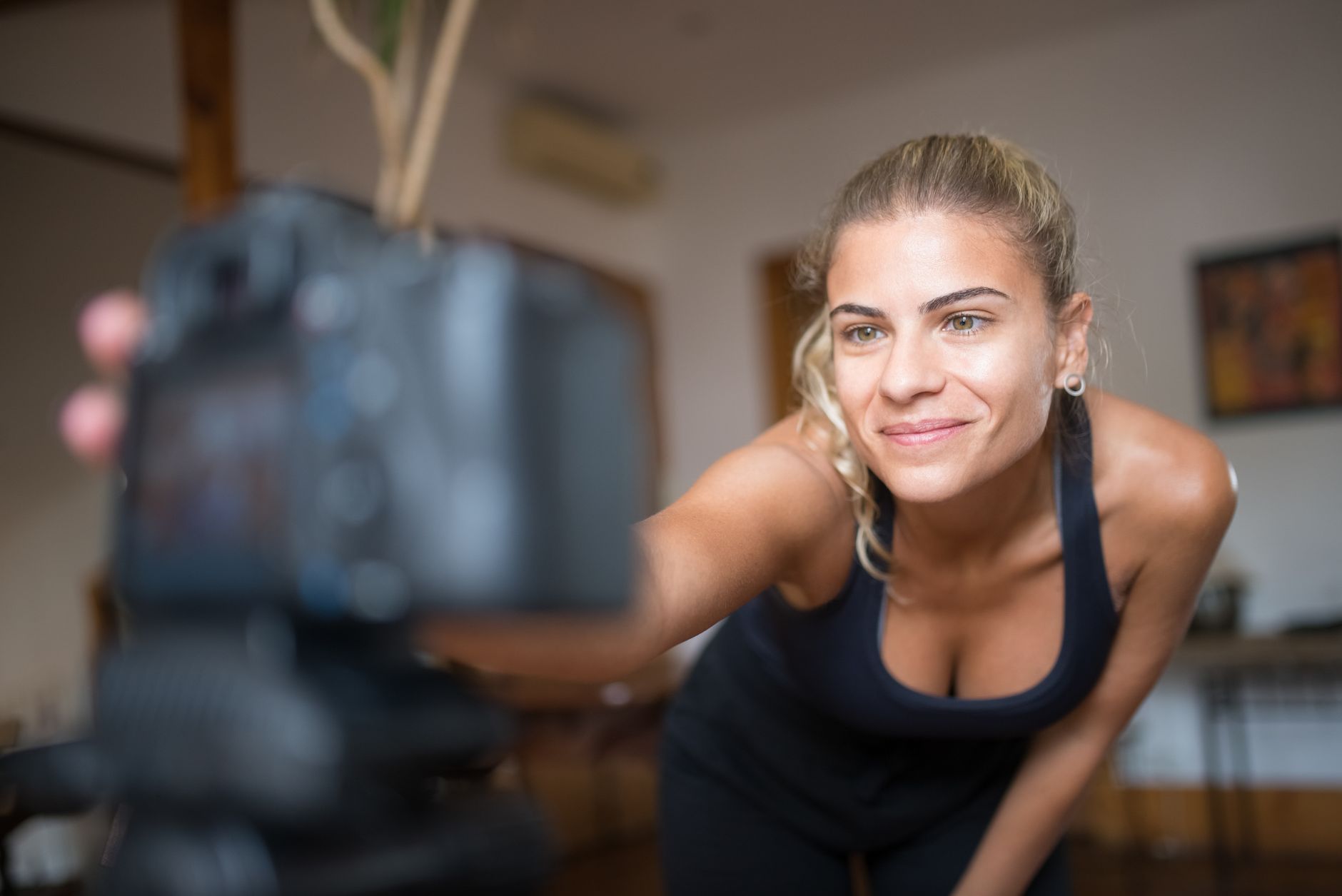 A smiling woman in sportswear leaning toward a camera set up on a tripod in a living room
